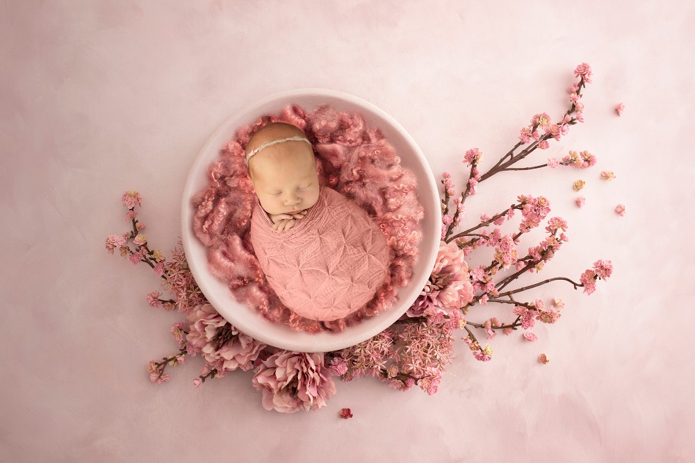 Newborn girl in pink wrap posed in white bowl with blooming pink flowers, dreamy Essex portrait